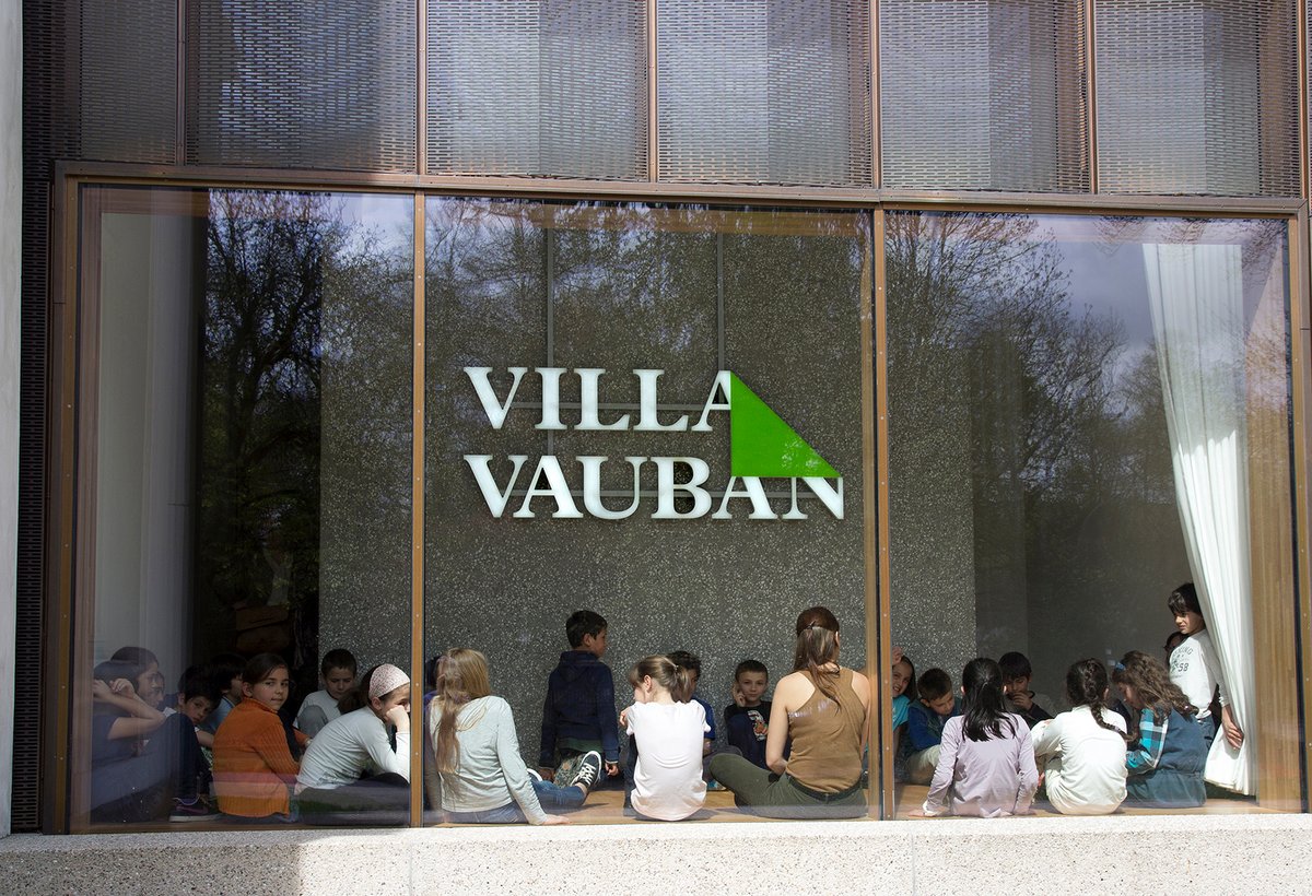 Children sitting behind a large window of the museum with a large logo of Villa Vauban in the background